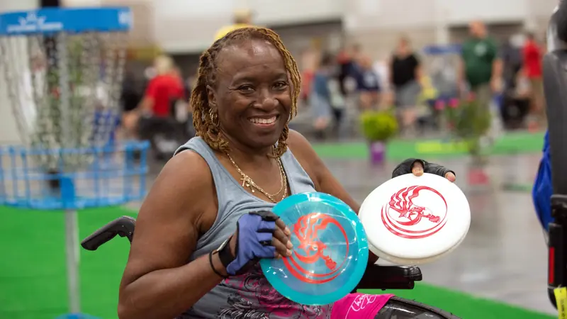 A smiling woman in a wheelchair holds two flying discs at an adaptive sports event